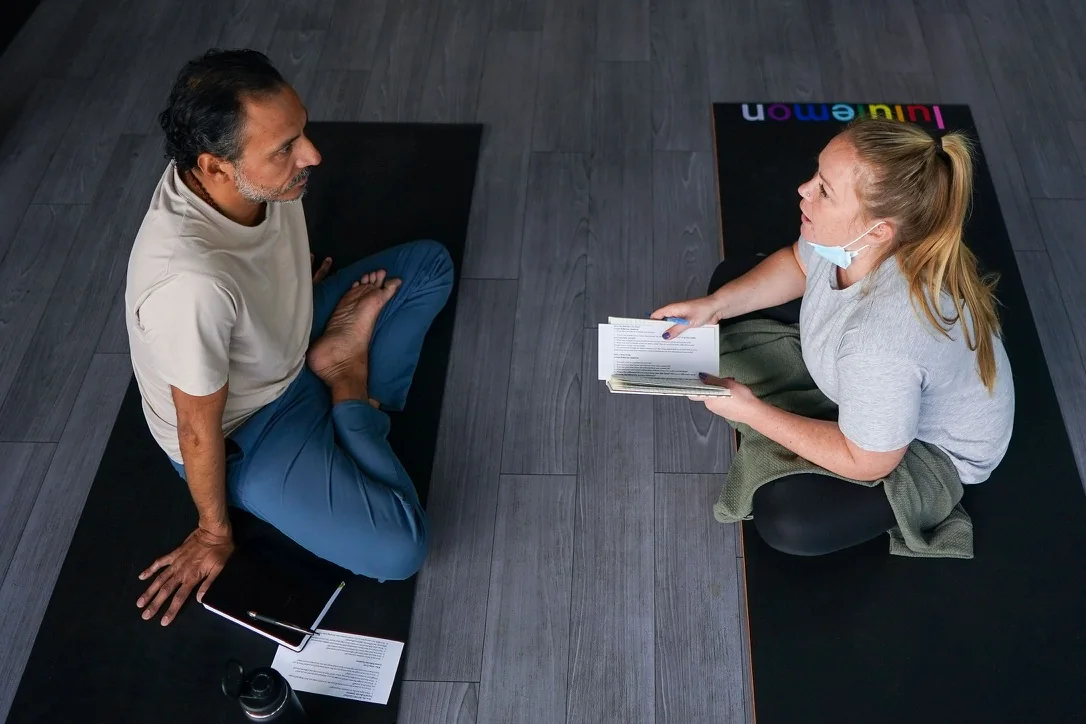 Jano Galindo seated on a mat in conversation with a teacher-training student holding notes.